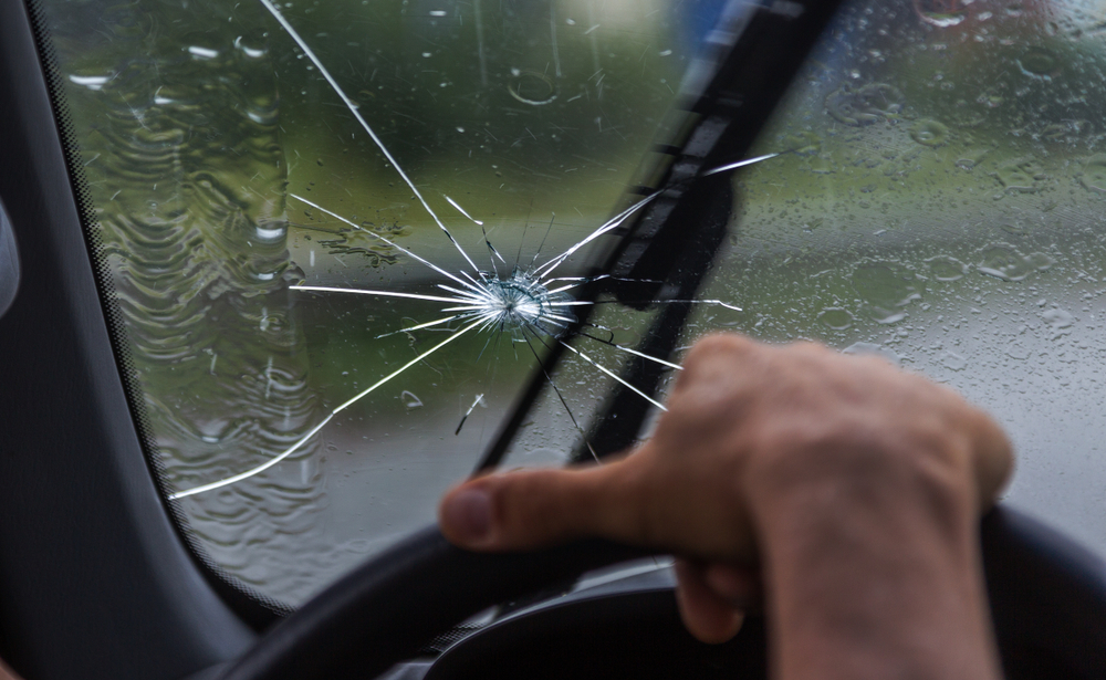 Man's hand on top of steering wheel with a windshield wiper scraping across a cracked windshield in the background